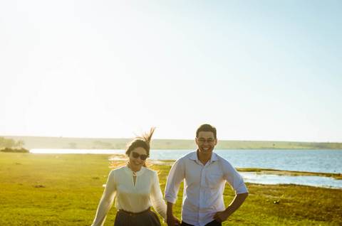 Ensaio fotográfico com um casal. Eles estão correndo de mãos dadas e sorrindo. Ao fundo tem o rio Tietê e está um dia ensolarado com céu azul claro.'