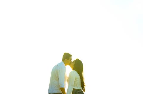 Foto com o casal se beijando de forma romântica, está com sol intenso e o fundo é amarelo bem claro.'