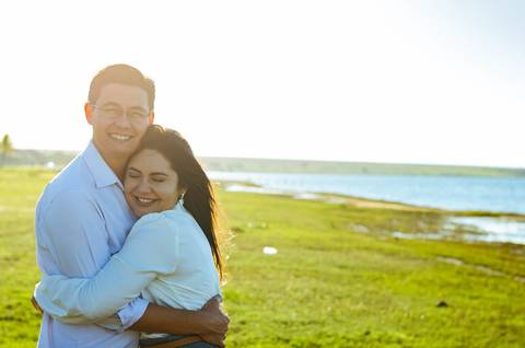 Fotografia de uma casal se abraçando com ternura. O dia tem sol forte, céu azul e um gramada bem verde.'