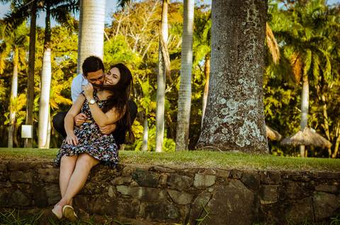 Fotografia de um casal. Ela está sentada em um pequeno muro de pedras. Ele chega por trás dela caminhando e a abraça, estão sorrindo e a vontade. Ao fundo há muitas palmeiras.'