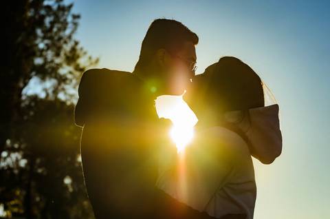 Fotografia silhueta de uma casal se beijando com ternura e carinho. Ao fundo tem o sol intenso.'