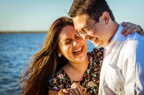 Foto do ensaio, o casal está dando gargalhadas juntos, ao fundo tem o rio Tietê e o céu azul.'