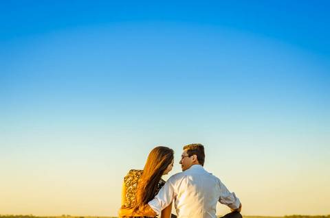 Fotografia do casal sentados, se abraçando e se olhando. A luz é do por do sol e ao fundo tem o céu bem azul. '