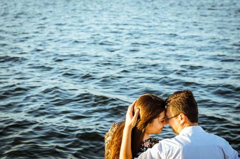 Fotografia do casal sentados, se abraçando e se olhando. A luz é do por do sol e ao fundo tem rio Tietê.'