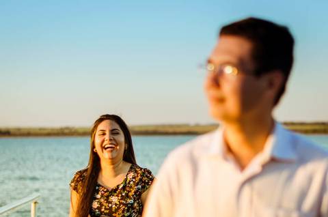 Fotografia do casal em um ensaio fotográfico. Ele está desfocado no primeiro plano e sua esposa está focada ao fundo olhando para ele e dando gargalhadas.'