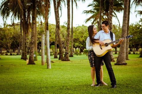 Foto no ensaio. Eles são casados. Ele está tocando violão e ela está por tráz dele o abraçando.. Estão em um lindo gramado e ao fundo várias plameiras.'