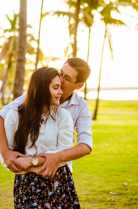 Foto do casal em um ensaio que estão se abraçando com ternura. ao fundo tem o gramada verde, o sol, o rio, e as palmeiras.'