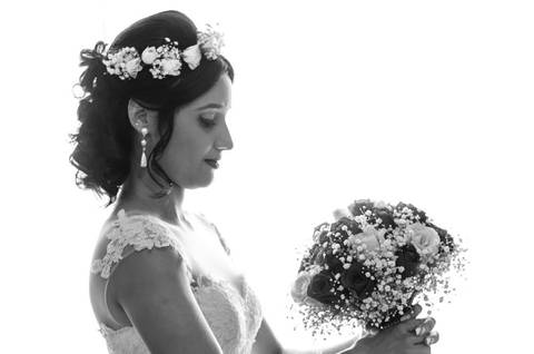 Fotografia em preto e branco de casamento do making of da noiva no salão de beleza no dia da noiva. O penteado no cabelo e maquiagem estão prontos, está de vestido de noiva branco longo segurando e olhando para o bouquê ou buquet. Em Araçatuba SP'