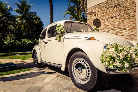 Fotografia de casamento com cerimônia e festa e recepção ao ar livre durante o dia no Califórnia Eventos em Araçatuba SP. Fusca branco com decoração de flores, céu azul e árvores de palmeiras ao fundo.'