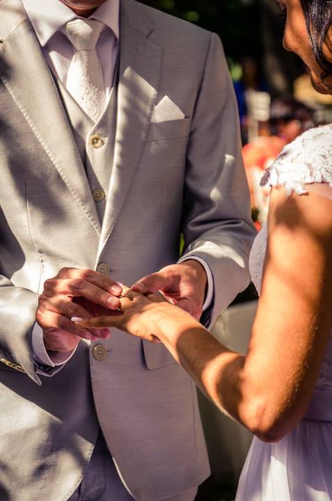 Fotografia de casamento com cerimônia e festa e recepção ao ar livre durante o dia no Califórnia Eventos em Araçatuba SP. Foco apenas nas mãos dos noivos. O Noivo de terno e colete cinza está colando as alianças na mão da noiva.'