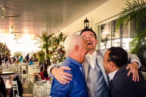 Fotografia de casamento com cerimônia e festa e recepção ao ar livre durante o dia no Califórnia Eventos em Araçatuba SP. O noivo abraça e sorri dois convidados. Ao fundo as mesas com decoração de flores.'