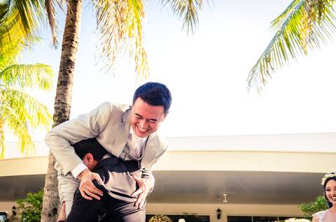 Fotografia de casamento com cerimônia e festa e recepção ao ar livre durante o dia no Califórnia Eventos em Araçatuba SP. Um amigo no noivo o segura nos ombros. O noivo de terno cinza sorri se divertindo. Ao fundo céu azul e palmeiras.'
