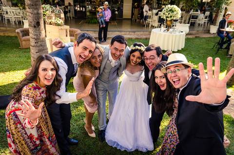 Fotografia de casamento com cerimônia e festa e recepção ao ar livre durante o dia no Califórnia Eventos em Araçatuba SP. Os noivos ao centro com os convidados e amigos, todos sorrindo se abraçando. Ao fundo as mesas do buffet e gramado verde.'