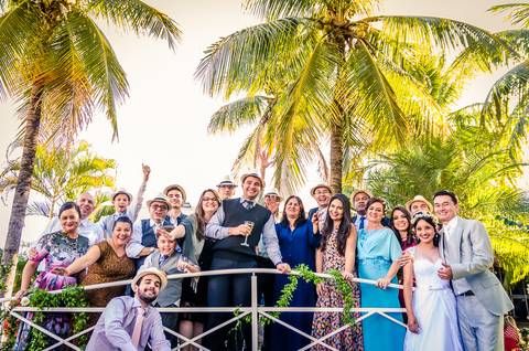 Fotografia de casamento com cerimônia e festa e recepção ao ar livre durante o dia no Califórnia Eventos em Araçatuba SP. Os noivos na lateral com os amigos sob uma pequena ponte com decoração de flores e ao fundo céu azul e árvores de palmeiras.'