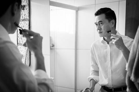 Fotografia de casamento em preto e branco do making of do noivo se olhando no espelho fazendo a barba na casa de seus pais, usando uma camisa branca. Em Araçatuba SP'