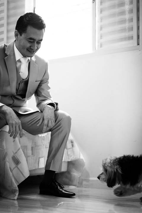 Fotografia de casamento em preto e brando do making of do noivo na casa de seus pais. Ele está de terno com colete e gravata tradicional sentado na cama brincando com seu cachorro que anda pelo quarto. Araçatuba, SP'