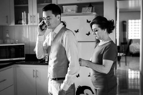 Fotografia de casamento em preto e branco do making of do noivo na casa de seus mais no dia do casamento. Ele veste camisa branca, colete e gravata e está ao celular na cozinha. Sua mãe está atrás dele ajudando ele a arrumar sua roupa. Em Araçatuba SP'