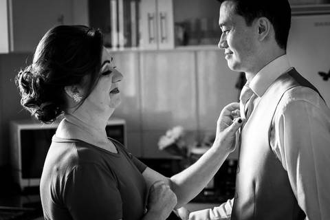 Fotografia de casamento em preto e branco do making of do noivo na casa de seus pais. Ele veste camisa branca, colete e gravata. Está frente para sua mãe que arruma sua gravata com expectativa e alegria. Em Araçatuba, SP'