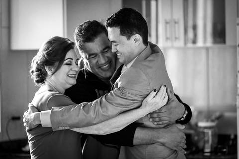 Fotografia de casamento em preto e branco do making of do noivo na casa de seus pais. Ele está pronto de terno, camisa, colete e gravata. Está abraçando seu pai e sua mãe com ternura. Araçatuba SP'