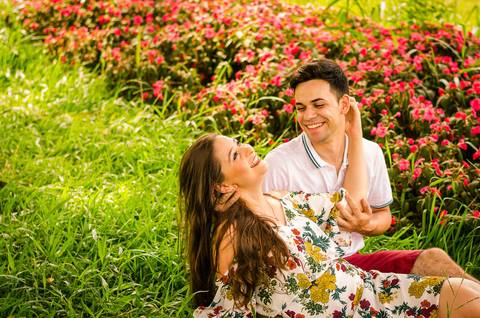 Fotografia do casal de noivos rindo juntos sentado no gramado e com flores vermelhas ao fundo em Maringá, PR'