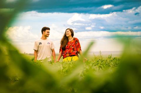 Fotografia do noivo e da noiva de frente e sorrindo em meio a uma plantação de soja com céu azul com algumas nuvens Maringá, PR'
