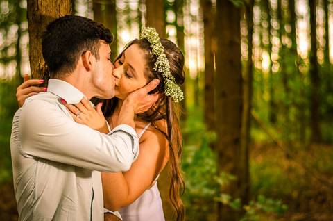 Foto do casal no pre casamento se beijando, ela com uma coroa de flores e vestido branco longo, ele com uma camisa clara de manga longa dobrada. Ao fundo uma floresta de árvores em Maringá, PR'