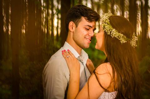 Foto do casal no pre casamento se beijando, ela com uma coroa de flores e vestido branco longo, ele com uma camisa clara de manga longa dobrada. Ao fundo uma floresta de árvores em Maringá, PR'