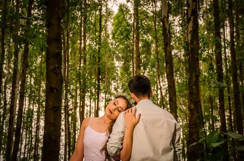 Foto do casal no pre casamento abraçados, ela encosta a cabeça com serenidade no ombro dele. ela com flores no cabelo e vestido branco longo, ele com uma camisa clara de manga longa dobrada. Ao fundo uma floresta de árvores em Maringá, PR'