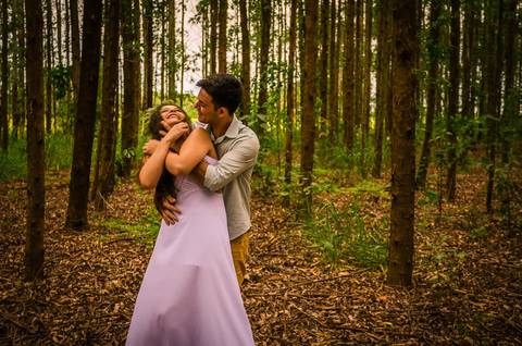 Foto do casal no pre casamento se abraçando e sorrindo um para o outro, ela usa flores no cabelo e vestido branco longo, ele com uma camisa clara de manga longa dobrada. Ao fundo uma floresta de árvores em Maringá, PR'