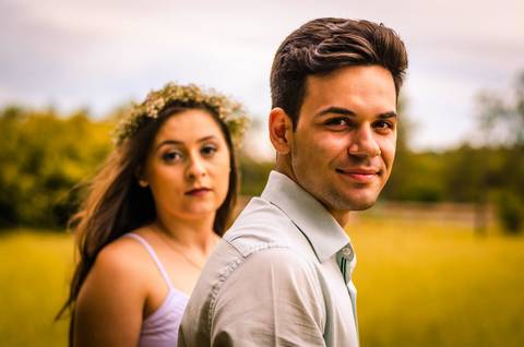 Fotografia do noivo em primeiro plano e da noiva em segundo plano, ambos com olhando para a câmera. Ela com flores no cabelo ao fundo tem um gramado em Maringá, PR '
