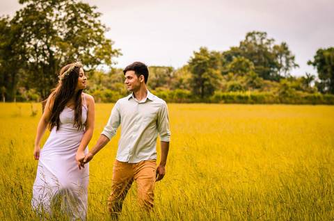 Fotografia do noivo e da noiva no pre casamento de mãos dadas no ensaio no campo se olhando e ela com flores no cabelo em Maringá, PR'