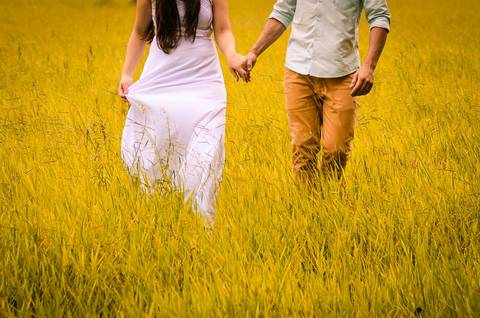 Fotografia do casal no pre casamento caminhando de mãos dadas em um gramado sem mostrar o rosto, ela usa um vestido longo branco e ele uma macisa clara e calça cáqui em Maringá, PR'