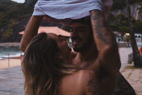 Matheus com os braços levantados colocando a camisa e Thais o abraçando. Os dois se olham olho no olho.'