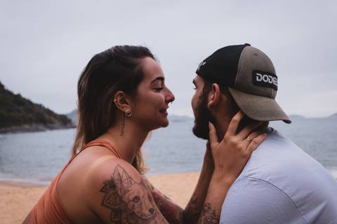 Retrato de Thais e Matheus sentados frente a frente. Matheus olha para o horizonte e Thais mantém seu olhar em Matheus. Praia vermelha, na urca, ao fundo.'