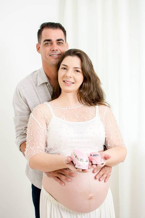uma gestante linda, de pele branca e cabelos castanhos esta abraçada com seu marido e segura em suas mãos dois carinhos em miniatura que representam suas filhas que vão nascer por nailie lucchesi e estudio amora'