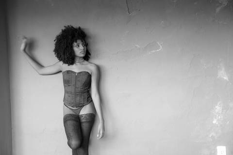 Ensaio sensual, onde a modelo usa um corpete e meia calça, encostada em uma parede.'