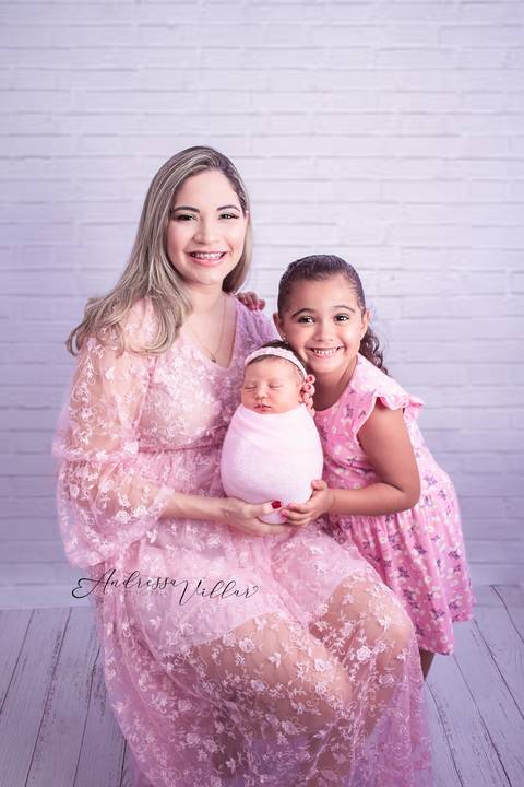 Mãe sentada de vestido rosa com a filha recém nascida enrolada no wrap rosa no colo e a filha mais velha de vestido rosa em pé ao lado'