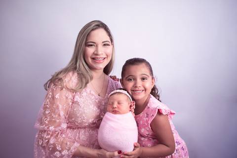 Mãe sentada de vestido rosa com a filha recém nascida enrolada no wrap rosa no colo e a filha mais velha de vestido rosa em pé ao lado'