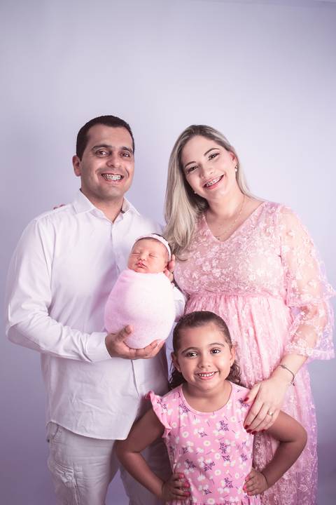 Pai de terno branco segurando a filha recém nascida enrolada no wrap rosa no colo mãe em pé ao lado do pai com filha mais velha em pé na sua frente, ambas de vestido rosa. '