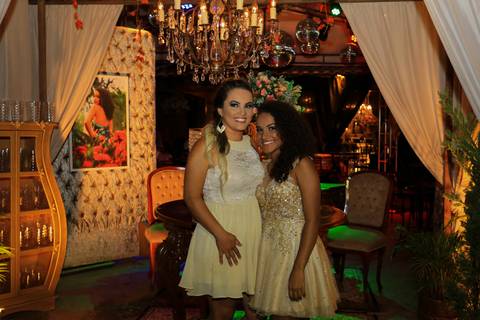 Aniversariante posa ao lado da mãe com decoração da festa ao fundo.'