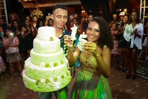 Hora do parabéns. Amigo segura o bolo, e aniversariante segura as velas, com grande sorriso'