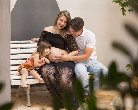 Família sentada no banco. Pai acaricia barriga da esposa gestante, enquanto irmã coloca ouvido na barriga da mãe'