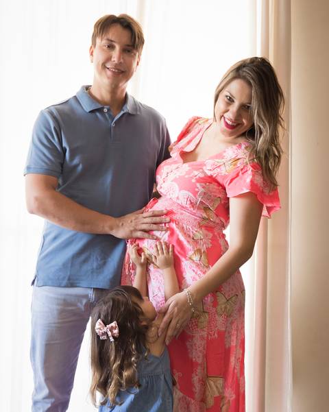 Família posa no contra luz da sala, acariciando a barriga. Pai e mãe olham para câmera, filha pequena acaricia barriga da mãe'