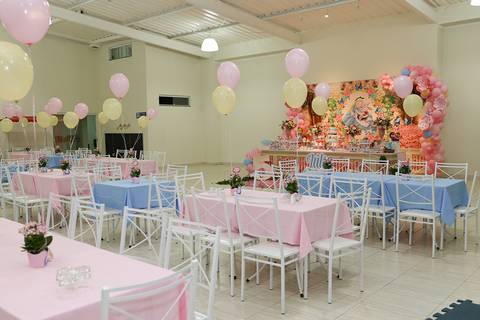 Visão geral do salão, mesa do bolo decorada ao fundo em tema Alice no País das Maravilhas e mesas decoradas com toalhas e balões'