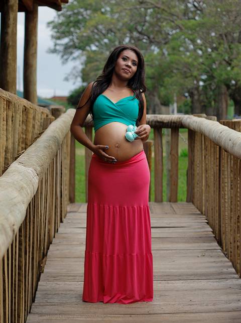 Gestante posa na casinha de madeira, olhando para o horizonte, acariciando a barriga e mostrando os sapatinhos do bebê'