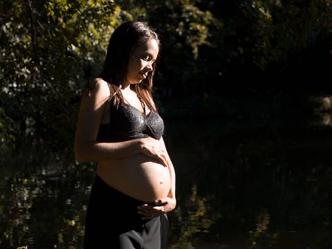 Gestante posa no parque, em frente ao lago, sob a luz do sol'