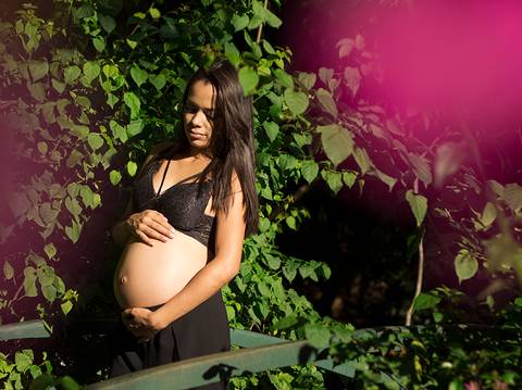 Gestante posa no parque, em meio a vegetação, sob a luz do sol, com flores rosas emoldurando a imagem'