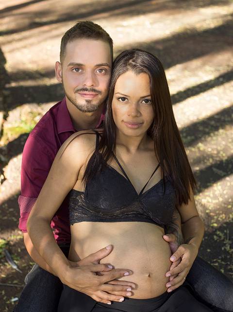 Casal posa sentado no banco de parque, posa olhando para câmera e acariciando a barriga'