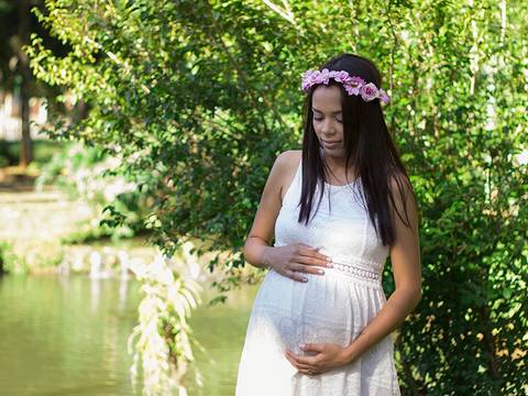 Gestante posa em frente a vegetação verde, com lago no fundo, de roupas brancas, coroa de flores, olhando para barriga e destacando a barriga'