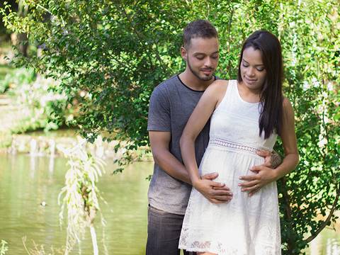 Gestante posa com pai do bebê, em frente a vegetação, com lago no fundo. Esposo abraça gestante a acaricia a barriga'